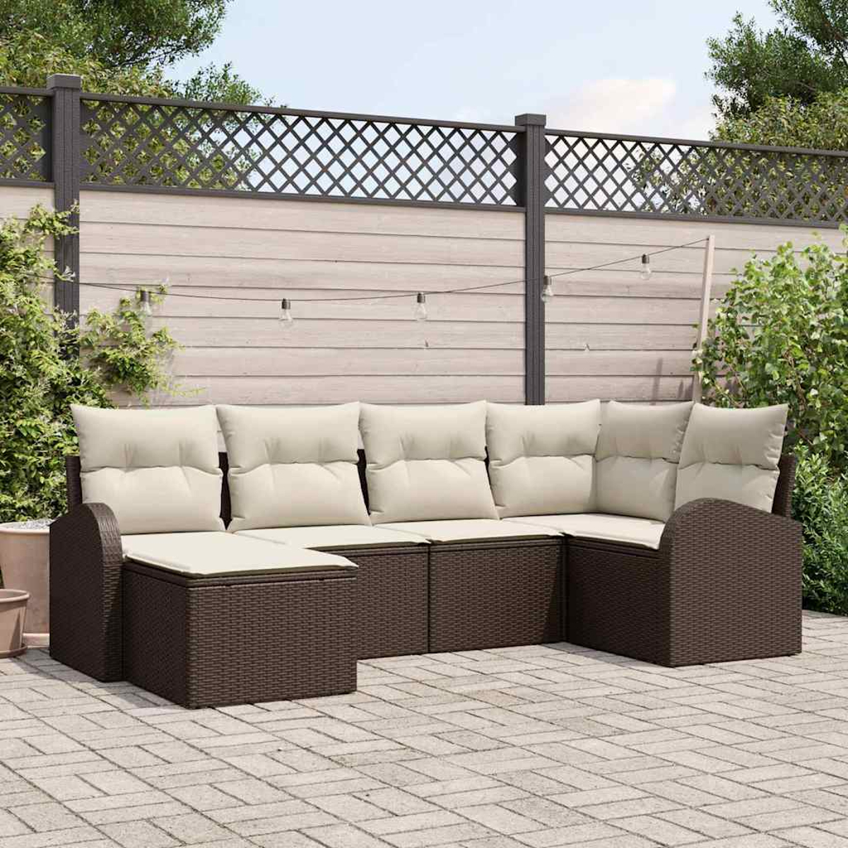 Outdoor-Ecksofa mit cremefarbenen Kissen. Brauner Korbrahmen. Dahinter ein Holzzaun mit Gitteraufsatz und Lichterketten.