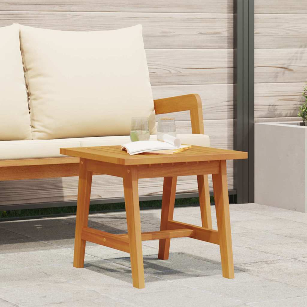 Una mesa auxiliar de madera con dos vasos y un libro en un patio. Un sofá con cojines crema está en el fondo.
