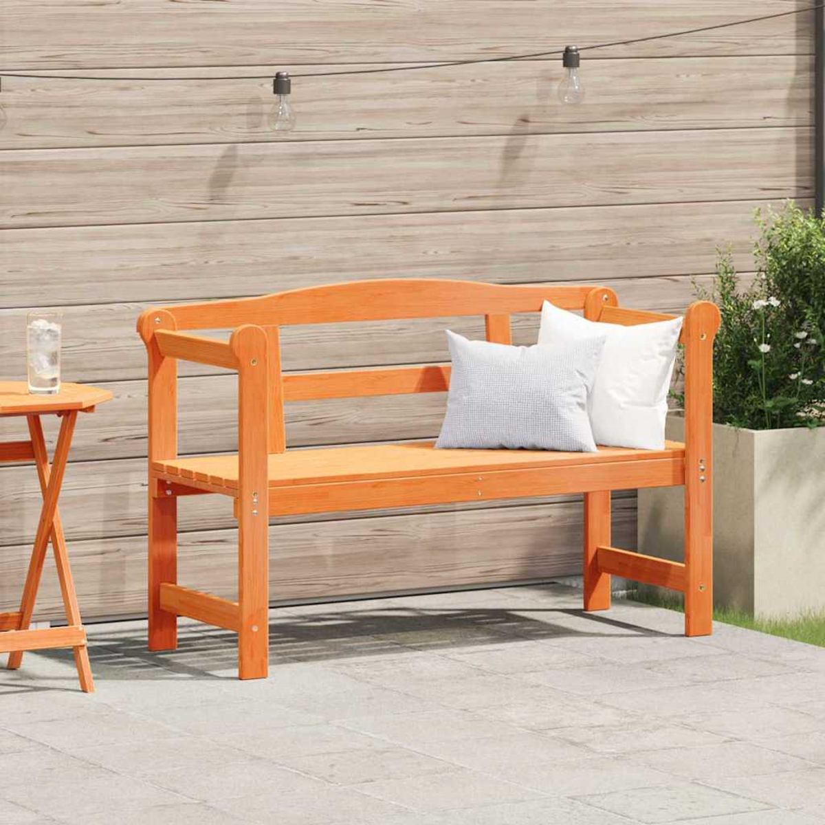 Banco de madera naranja con dos almohadas. A la izquierda, una mesita con un vaso de agua. El fondo es una pared de madera.