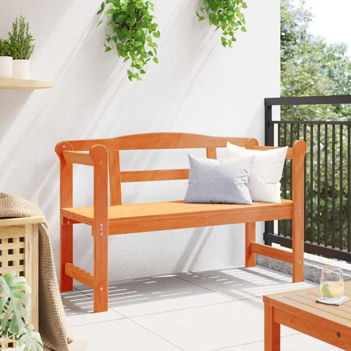 Banqueta de madera en balcón con almohadas. Plantas verdes cuelgan de la pared blanca. Mesa con vaso y barandilla al fondo.