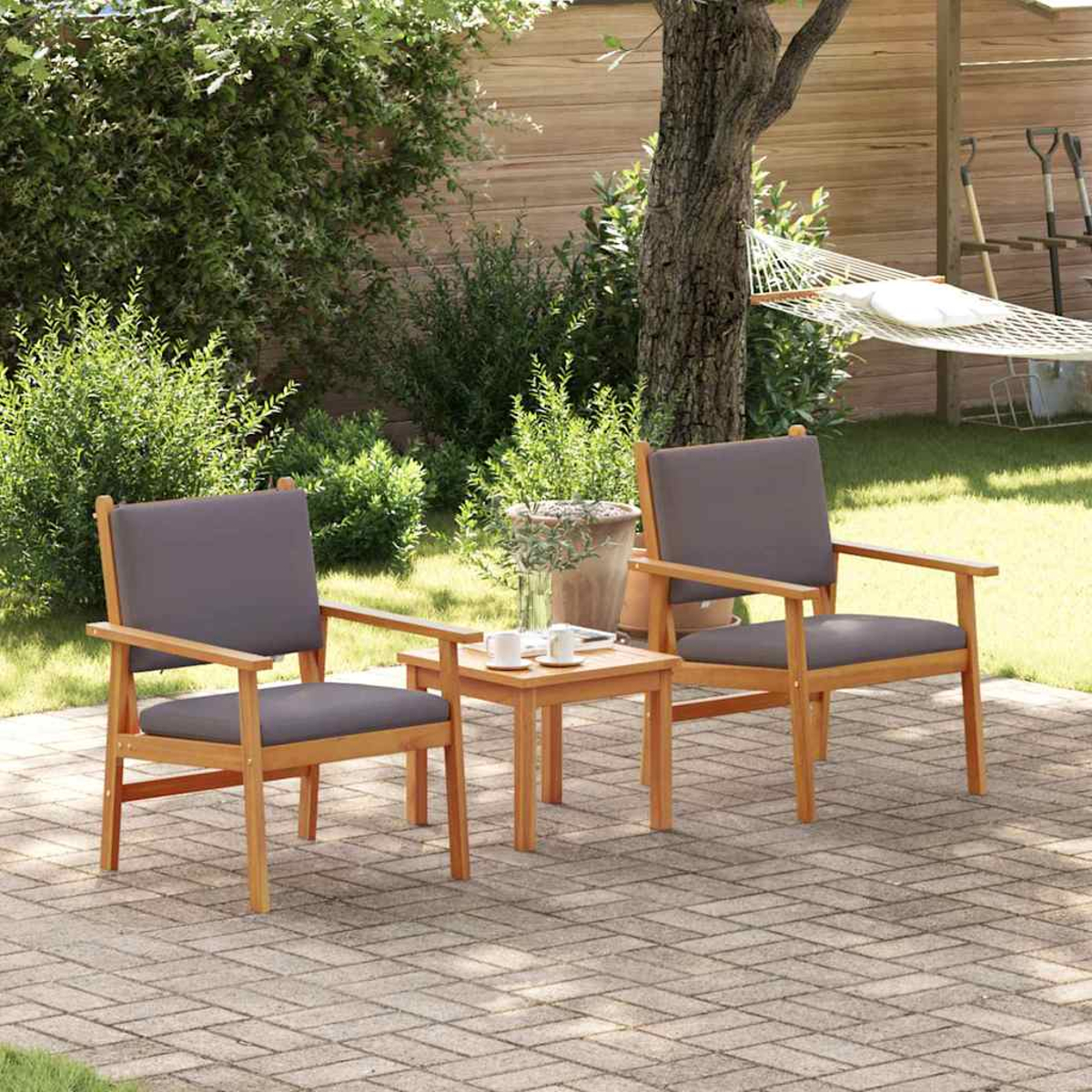 Zwei Holzstühle mit grauen Kissen und ein kleiner Tisch auf einer Ziegelterrasse. Im Hintergrund hängt eine Hängematte, üppiges Grün umgibt sie.