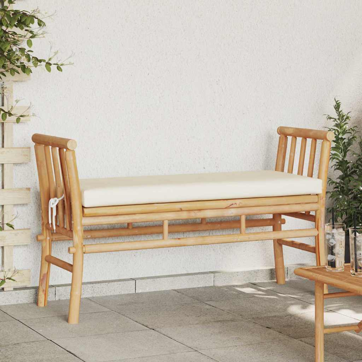 Eine Bambusbank mit cremefarbenem Kissen steht auf einer Terrasse. Ein kleiner Tisch mit Gläsern ist in der Nähe. Grünpflanzen befinden sich an den Seiten und oben links.