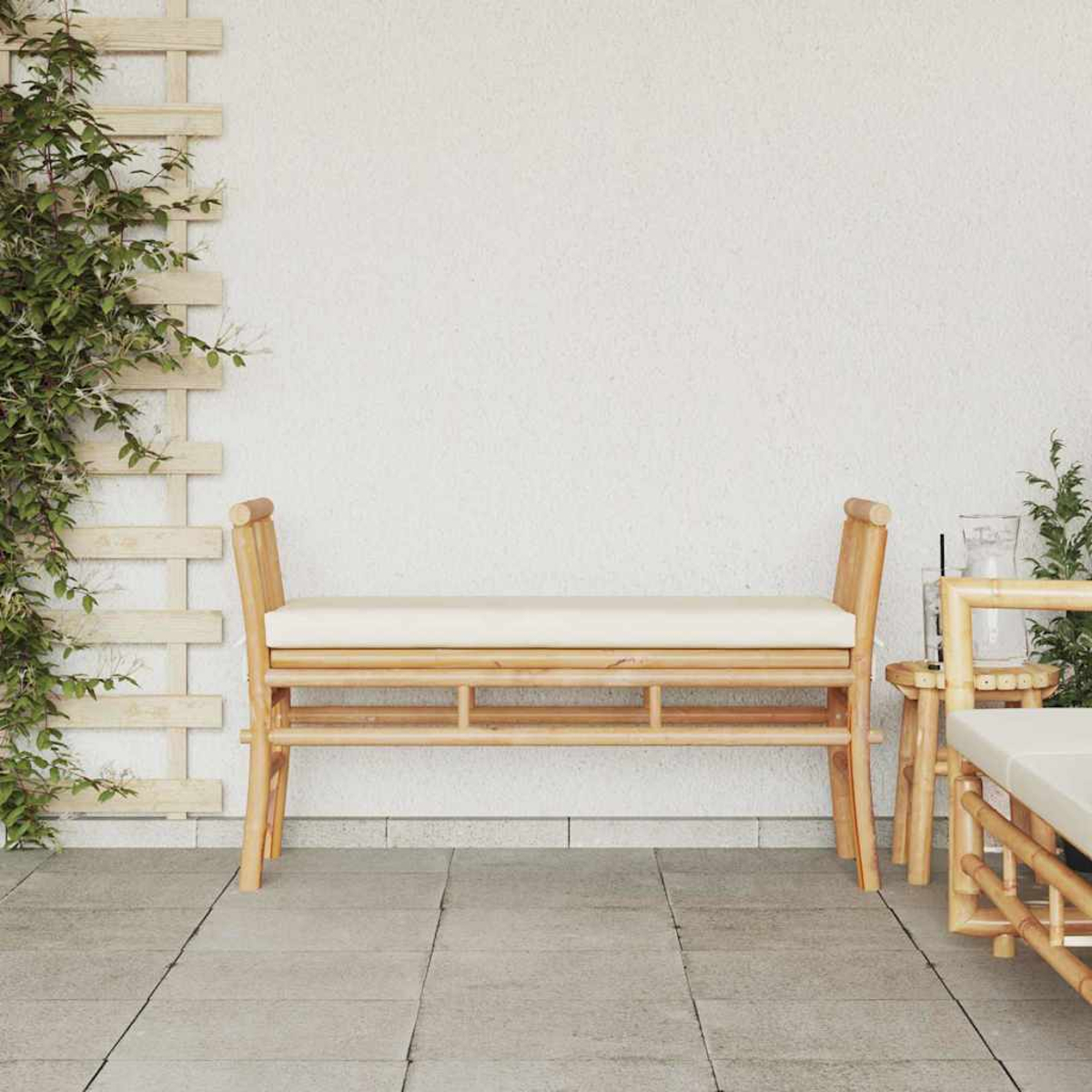 Eine Bambusbank mit weißem Kissen steht auf einer gefliesten Terrasse. Links ein Holzgitter mit grünen Weinranken. Im Hintergrund eine weiße Wand.