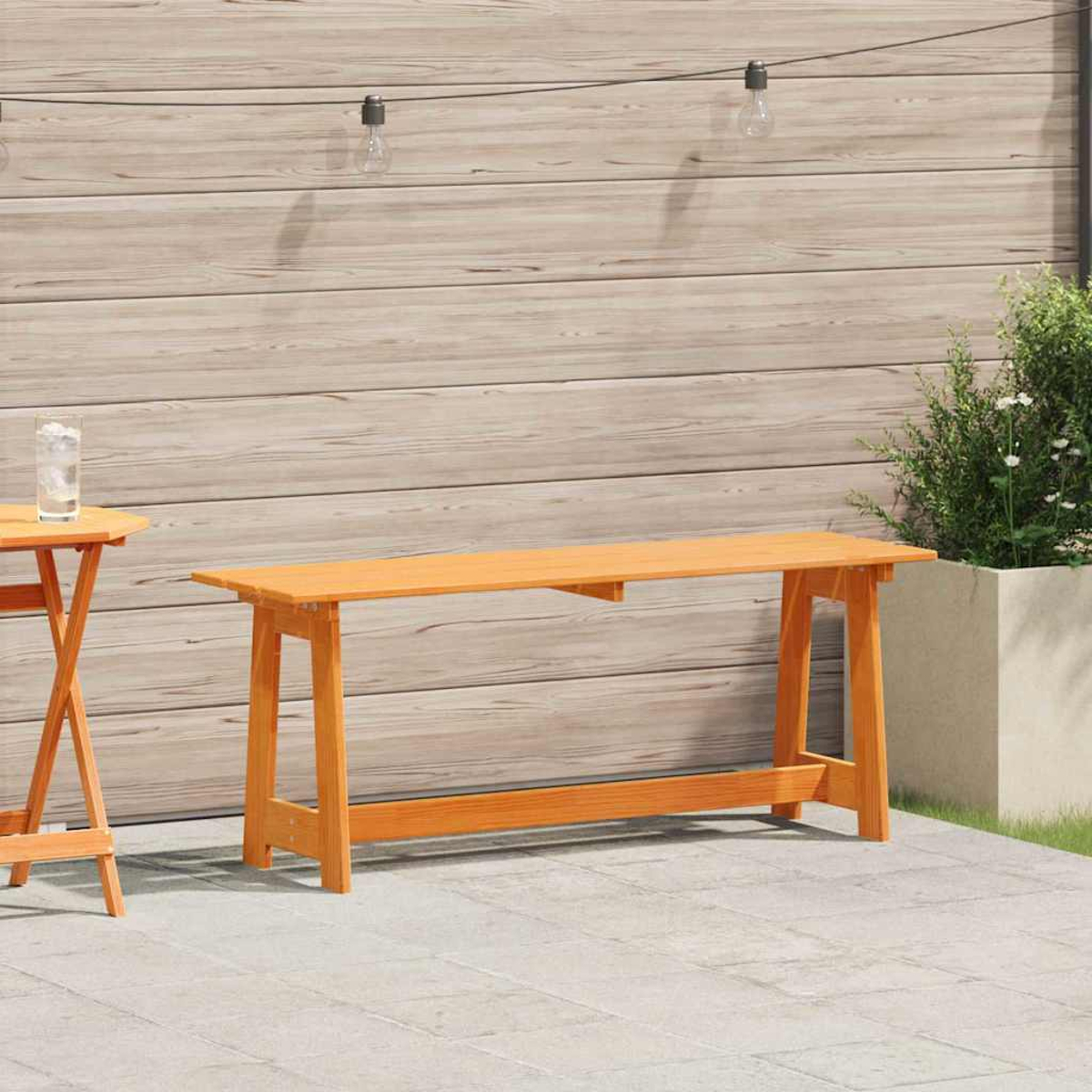 Eine Szene im Freien mit einer langen, orangefarbenen Bank und einem kleinen Tisch. Im Hintergrund eine Holzwand und Pflanzen.