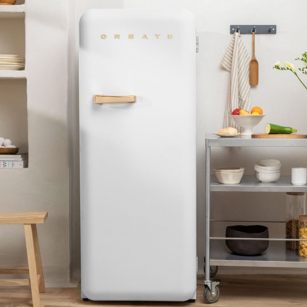 Un refrigerador blanco con 'CREATE' en letras doradas. Un tirador de madera y un carrito metálico con comida y cuencos a su lado.