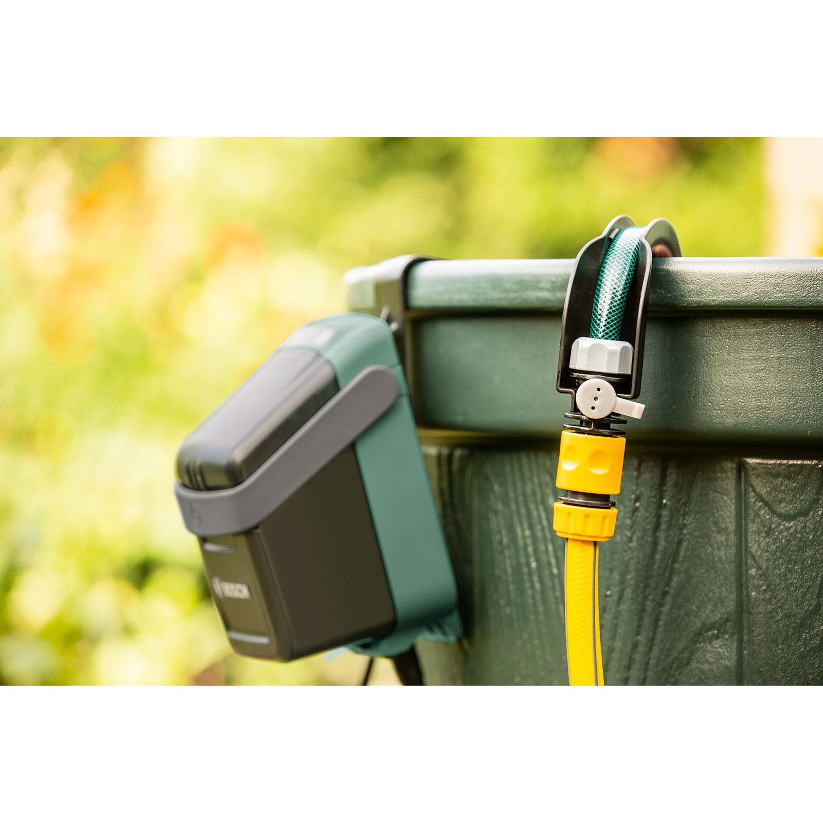 Zielony wąż ogrodowy i timer wody są przymocowane do zielonego zbiornika na wodę. Tło jest rozmyte.