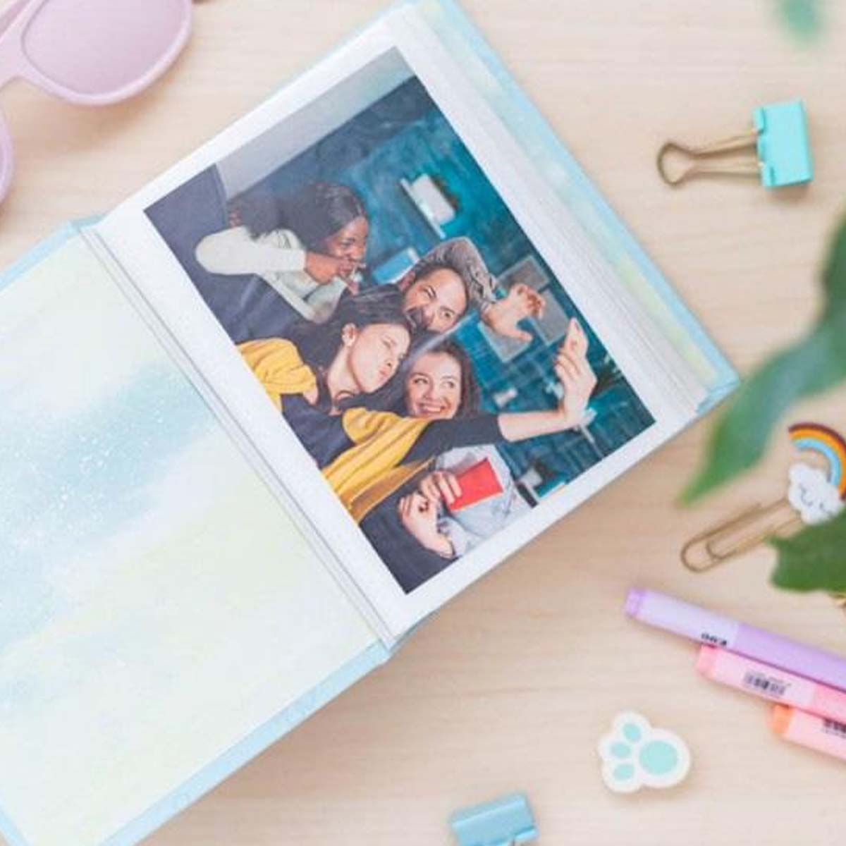 Un album photo montre quatre personnes prenant un selfie. L'album est sur une table en bois avec des articles de papeterie et des accessoires.