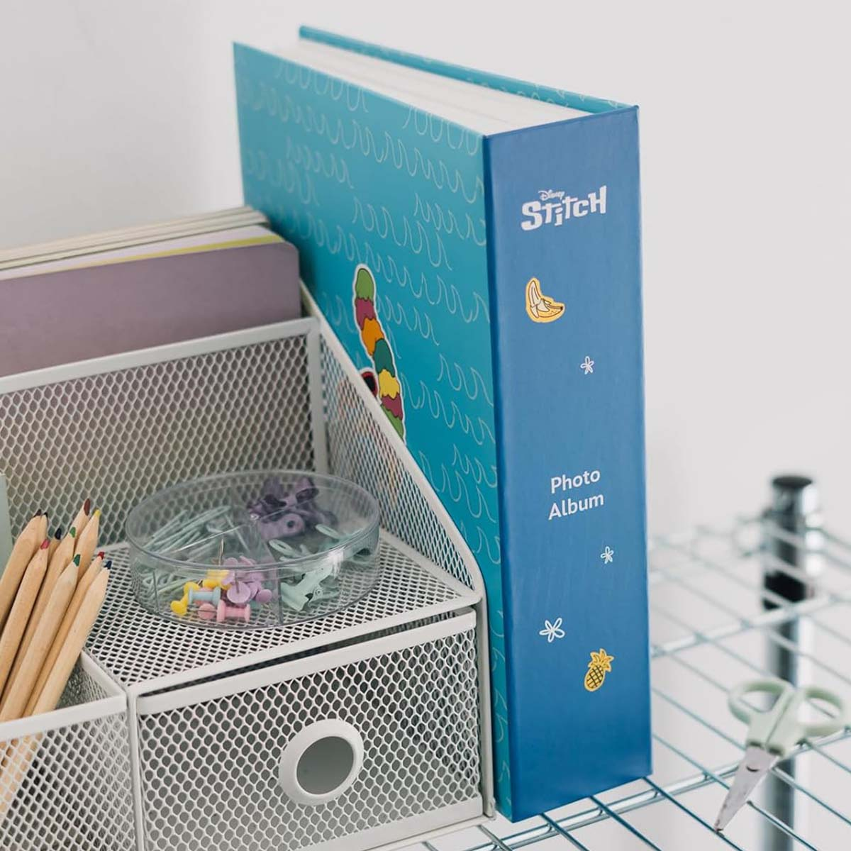 Ein blaues Fotoalbum mit Stitch-Design steht neben einem weißen Metall-Organizer mit Bleistiften, Pins und einer lila Ordner.