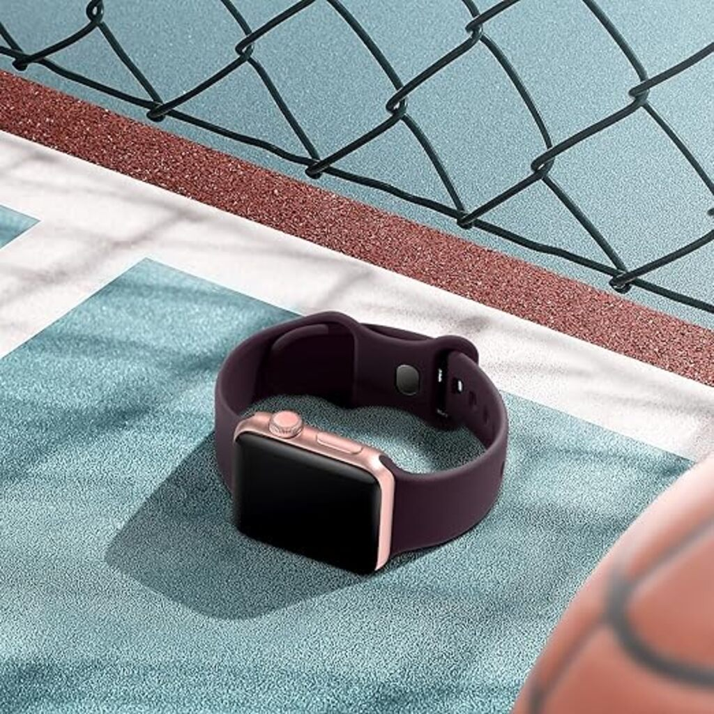 Eine Smartwatch mit dunkelviolettem Armband und roségoldenem Gehäuse liegt auf einer teal-farbenen Oberfläche. Im Hintergrund ist ein Drahtzaun.