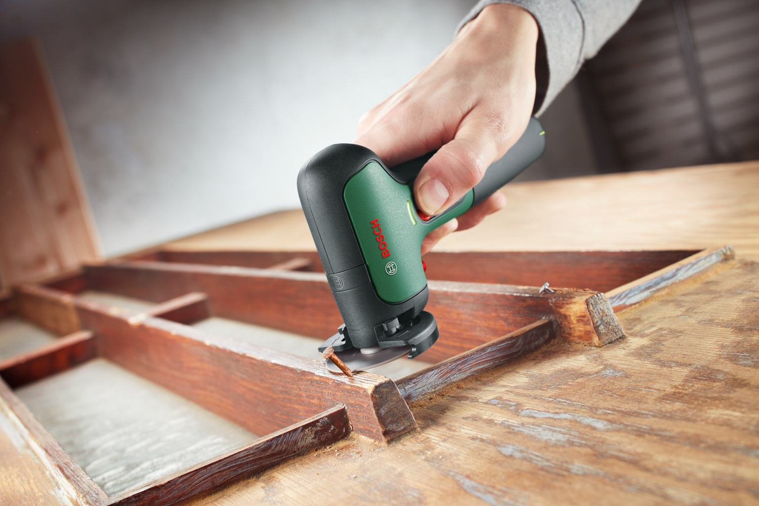 Une main tient un outil multifonction Bosch coupant du bois. L'outil est vert et noir. Le cadre en bois est marron, le fond est flou.
