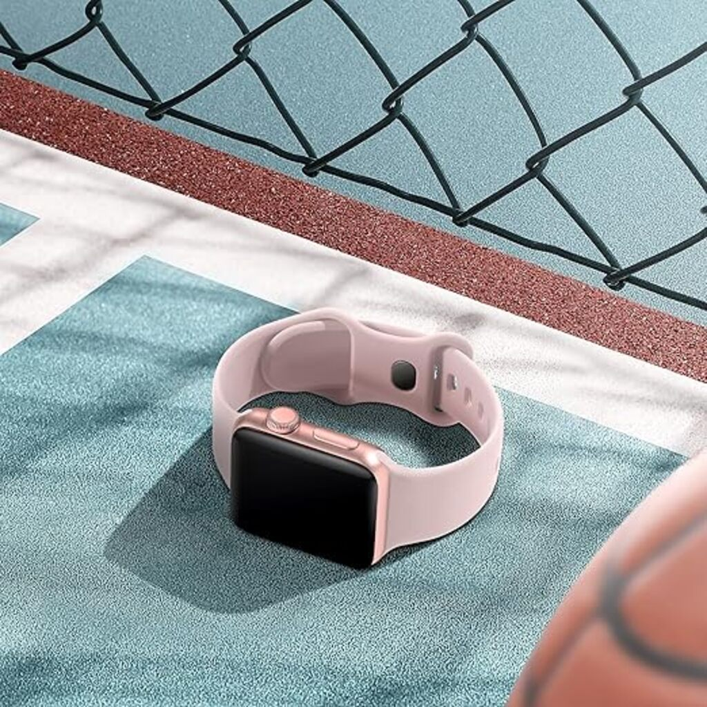 Eine rosa Smartwatch mit schwarzem Bildschirm liegt auf einer blauen Oberfläche, mit einem Zaun und Basketball im Hintergrund.