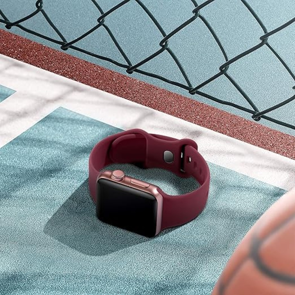 Eine weinrote Smartwatch liegt auf einer strukturierten blauen Oberfläche. Im Hintergrund befindet sich ein schwarzer Zaun.