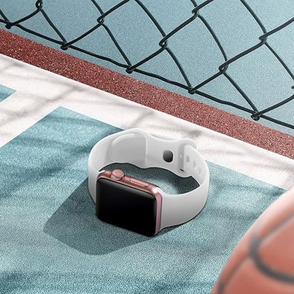 Eine weiße Smartwatch mit roségoldenem Gehäuse liegt auf einer blauen Oberfläche. Ein Drahtzaun und ein Basketball sind ebenfalls zu sehen.