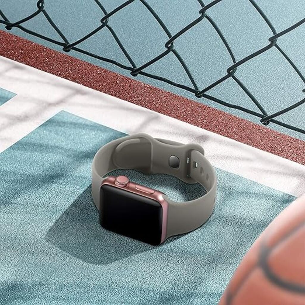 Eine roségoldene Smartwatch mit grauem Armband liegt auf einer blauen Oberfläche, mit einem Basketball in der Ecke und einem Zaun im Hintergrund.