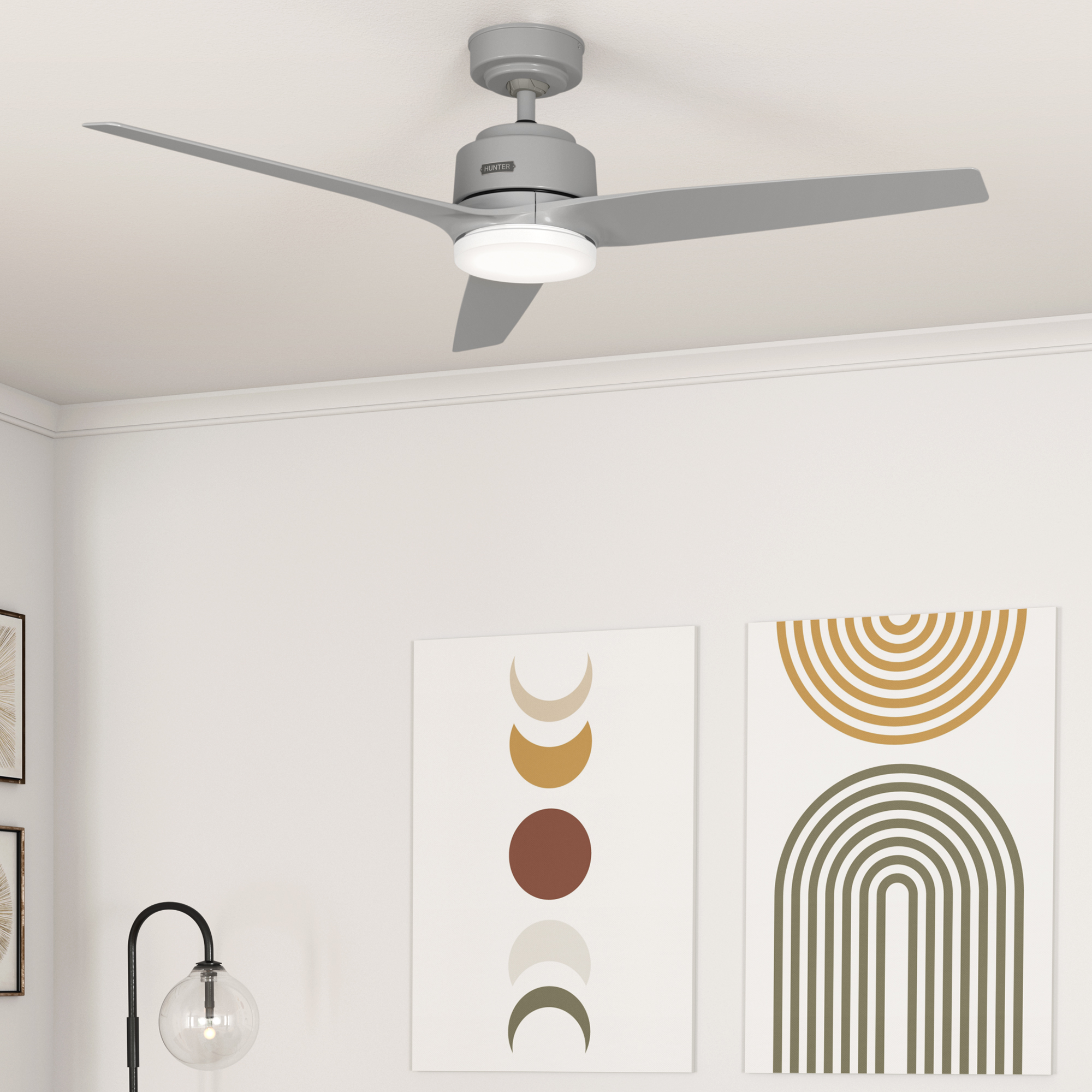Ein grauer Deckenventilator mit drei Flügeln ist an einer weißen Decke montiert. Darunter befinden sich zwei abstrakte Kunstwerke und eine Stehlampe.