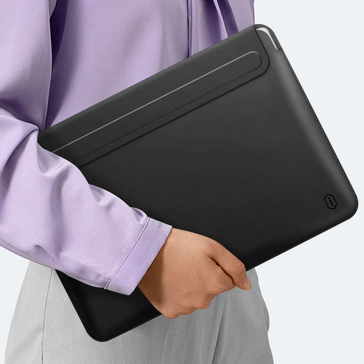 Une personne tient une housse d'ordinateur portable noire. Elle porte une chemise lilas et un pantalon gris. La housse a un petit logo.