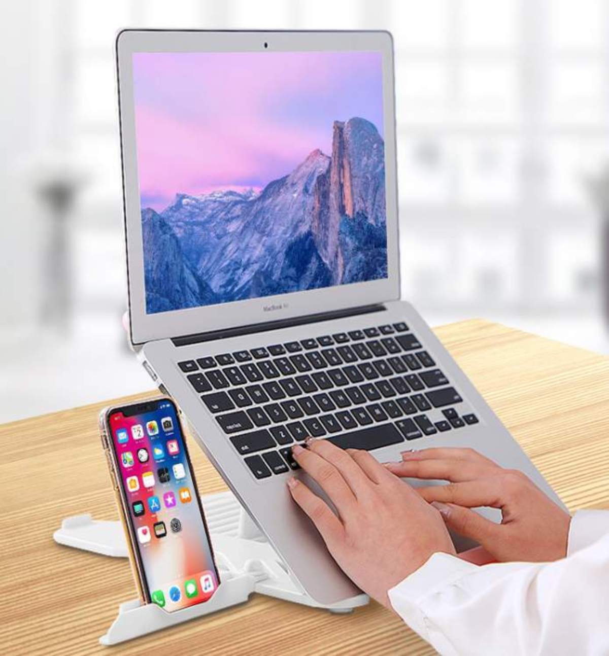 Laptop auf weißem Ständer mit Telefon. Der Laptop-Bildschirm zeigt ein Berg-Bild. Hände tippen auf dem Laptop. Holztisch im Vordergrund.