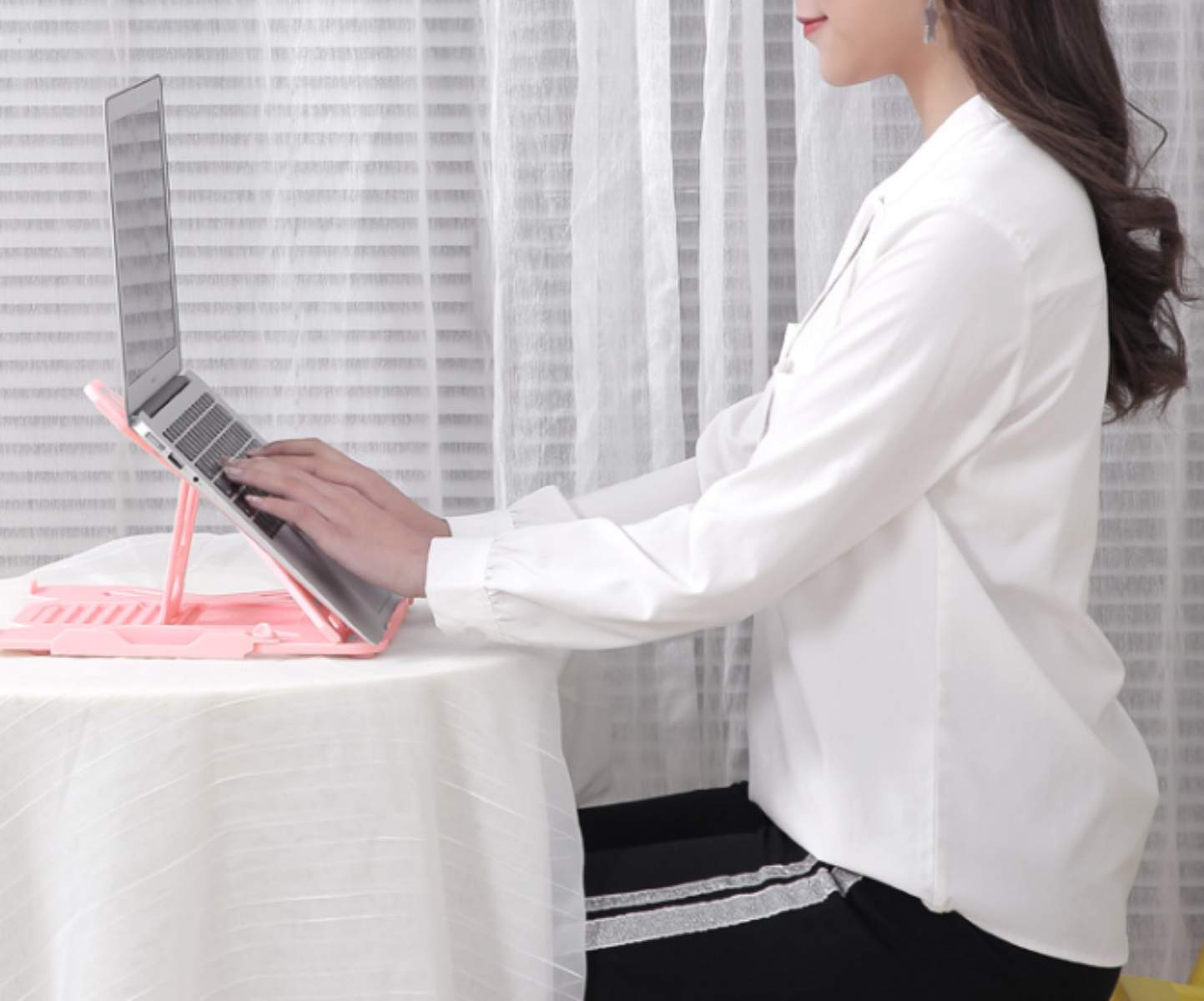 Frau benutzt Laptop auf rosa Ständer. Sie trägt ein weißes Hemd und schwarze Hose. Der Laptop steht auf einem Tisch.