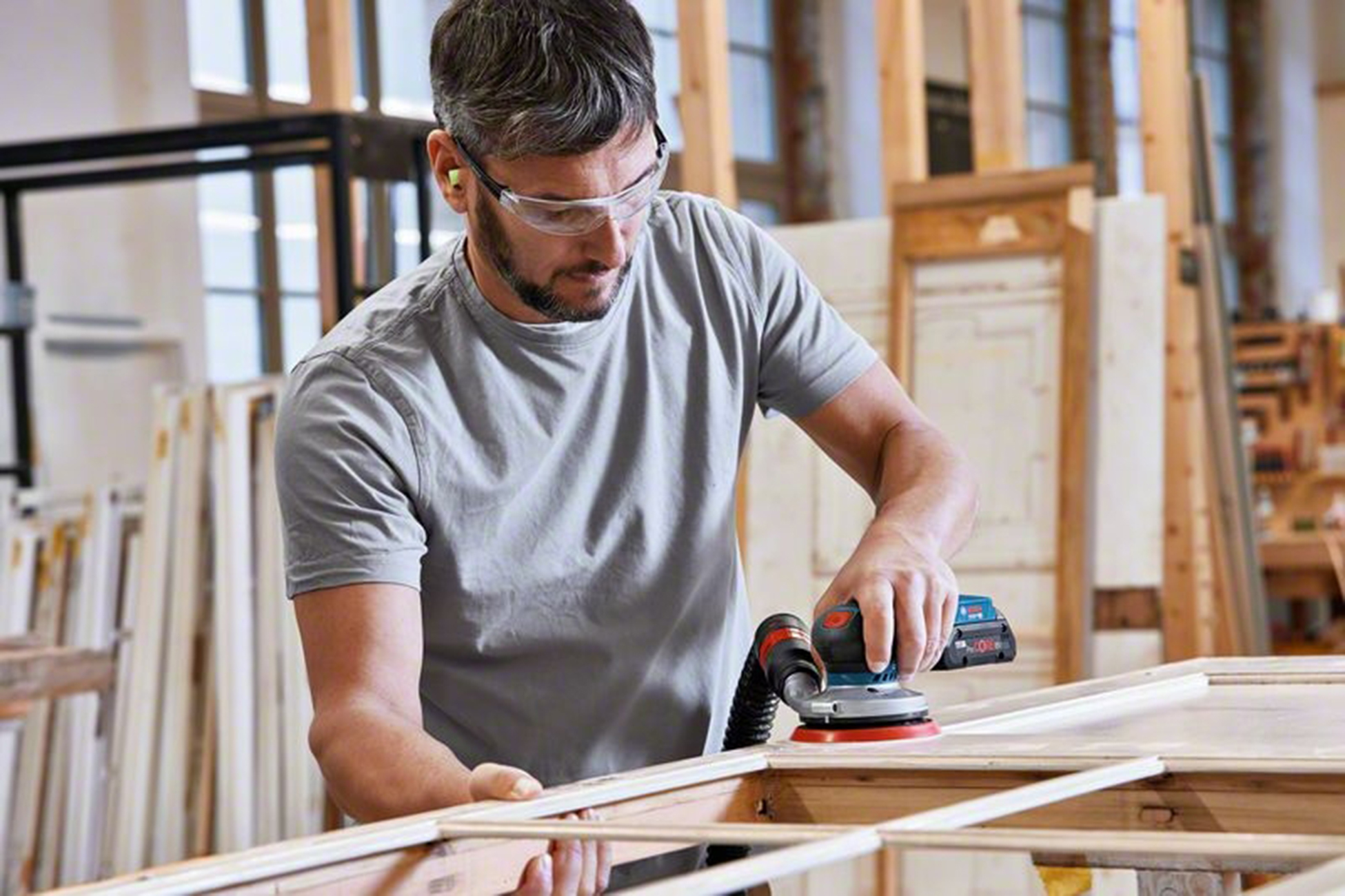 Mężczyzna szlifuje drewnianą ramę szlifierką elektryczną. Ma ochronę oczu i zatyczki do uszu. W warsztacie są drewniane ramy.