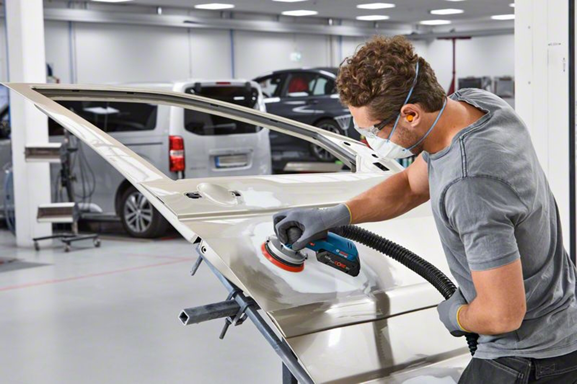 Mężczyzna szlifuje drzwi samochodu szlifierką. Nosi okulary ochronne, maskę i rękawice. W tle widać inne samochody.