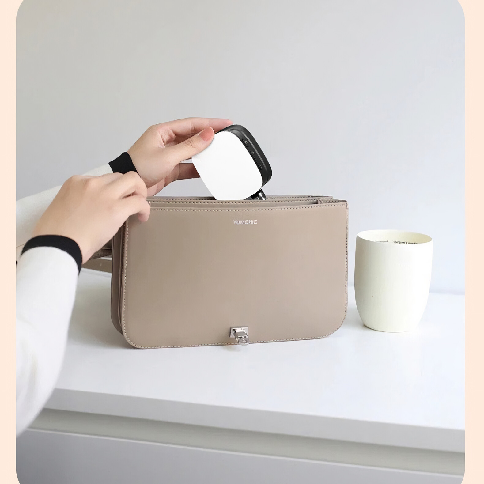 Eine Person legt ein weißes und schwarzes Gerät in eine beige Handtasche. Neben der Handtasche steht ein weißer Becher.