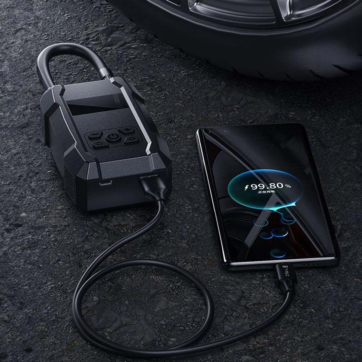 Schwarzes Gerät lädt ein Telefon auf. Telefon zeigt einen Ladestatus an. Autoreifen im Hintergrund. Beide Geräte sind mit einem schwarzen Kabel verbunden.