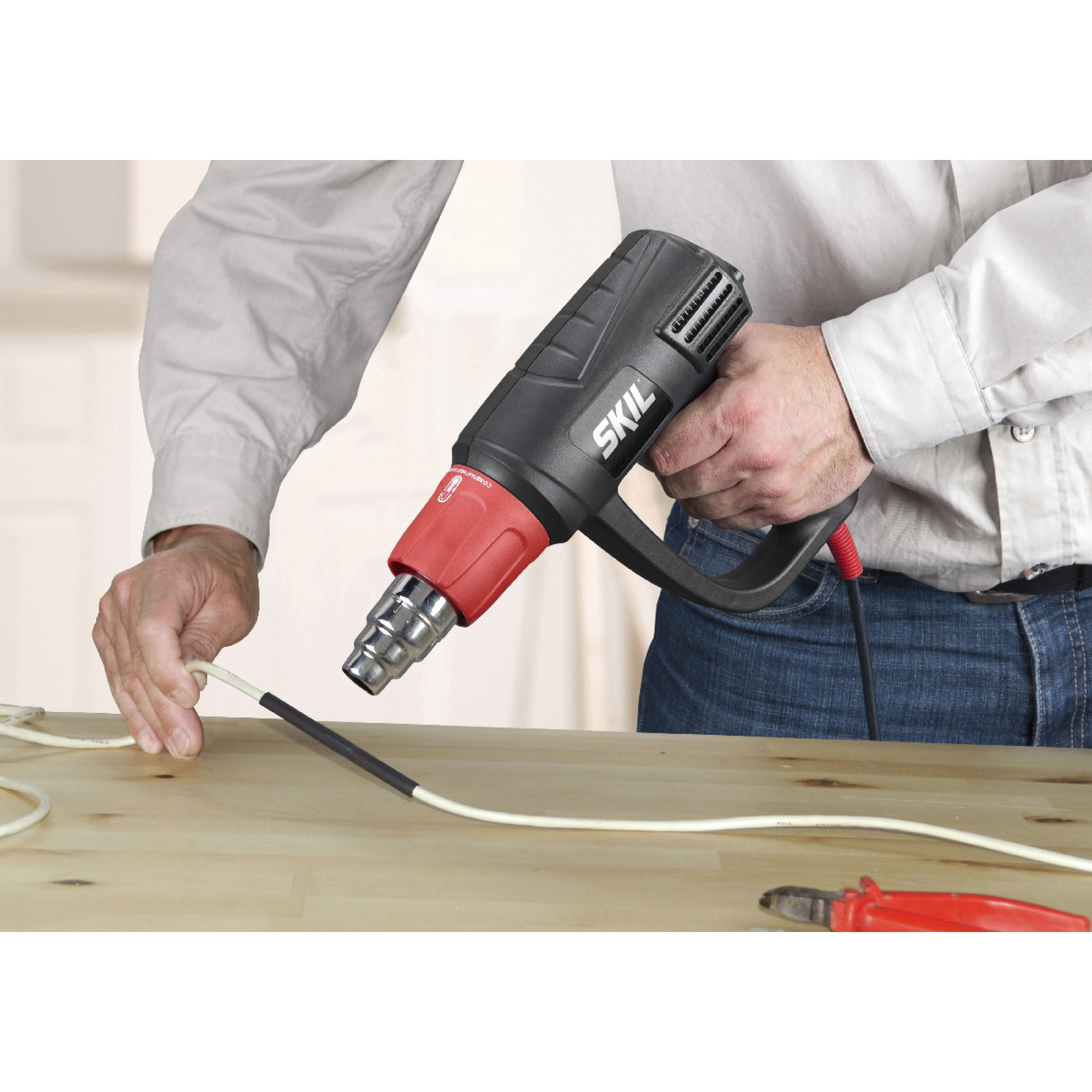 Een man gebruikt een heteluchtpistool op een draad. De tool is zwart en rood. De achtergrond is een houten tafel en een vage achtergrond.