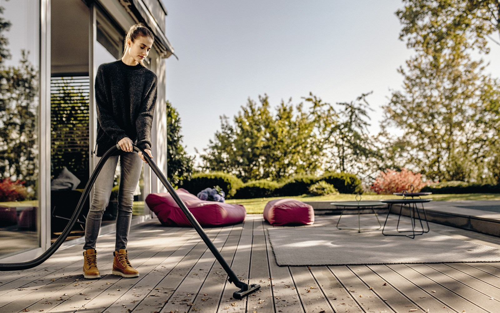 Kobieta odkurza drewniany taras. Ma na sobie sweter i dżinsy. W tle worki do siedzenia i drzewa.