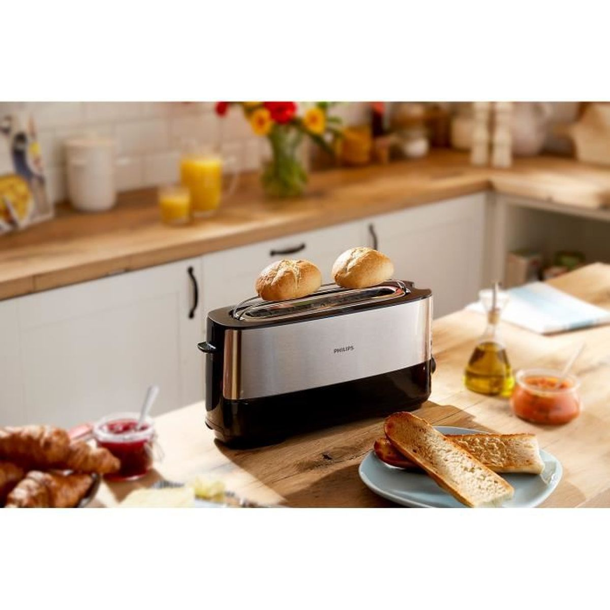 Ein Toaster mit zwei Brötchen oben drauf. Aufgeschnittenes Brot auf einem Teller. Croissants und Marmelade auf dem Tisch. Orangensaft und Blumen im Hintergrund.