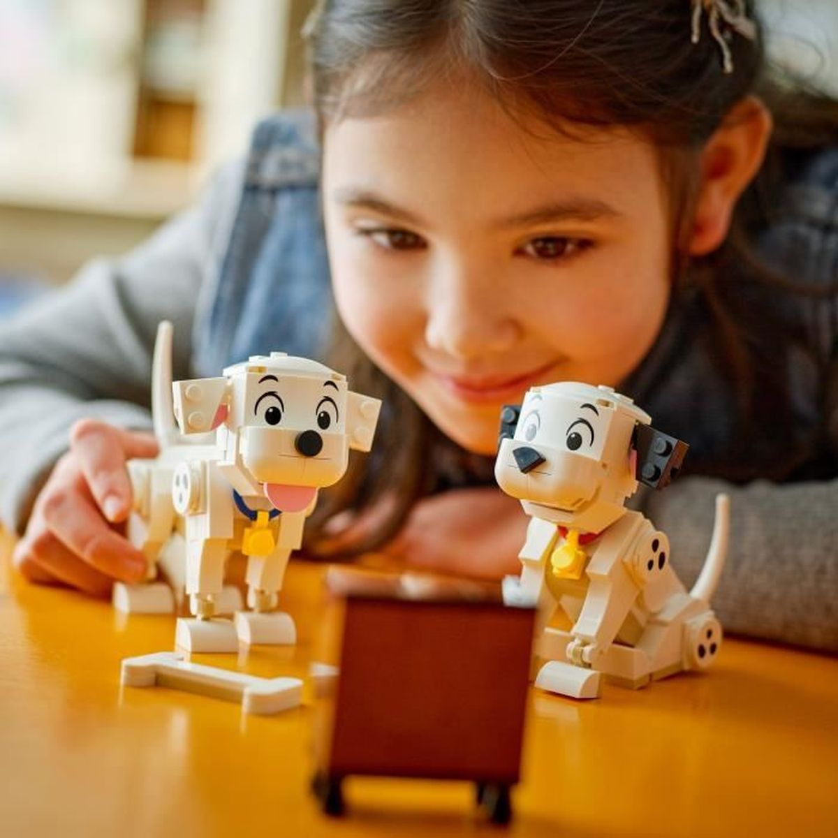 Bir kız, sarı bir masa üzerinde iki Dalmatian Lego oyuncağına gülümsüyor. Bir oyuncağın dili dışarıda, diğerinin kırmızı bir tasması var.