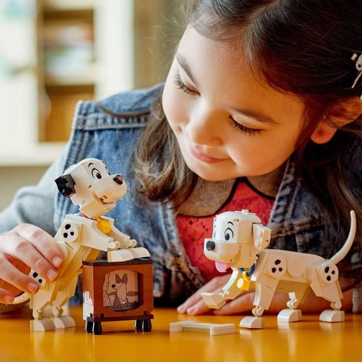 Bir kız Lego Dalmaçyalı figürleriyle oynuyor. İki köpek, biri TV'de ve üçüncüsü yerde. Kız onlara bakıyor.