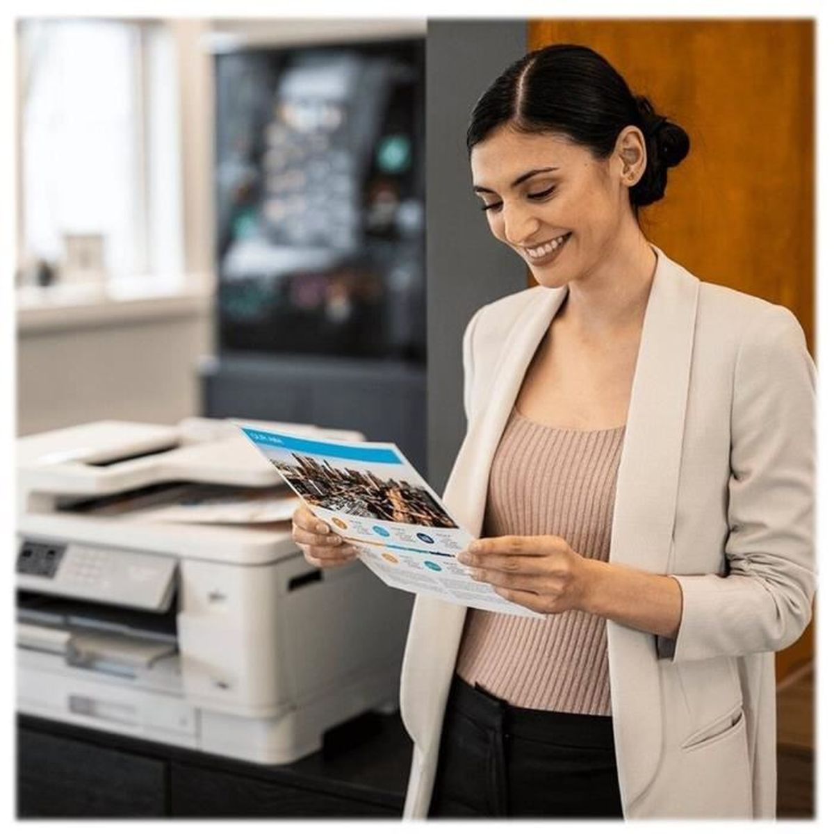 Kobieta w marynarce uśmiecha się, patrząc na wydrukowany dokument. Po lewej stronie drukarka, pod nią ciemna szafka.