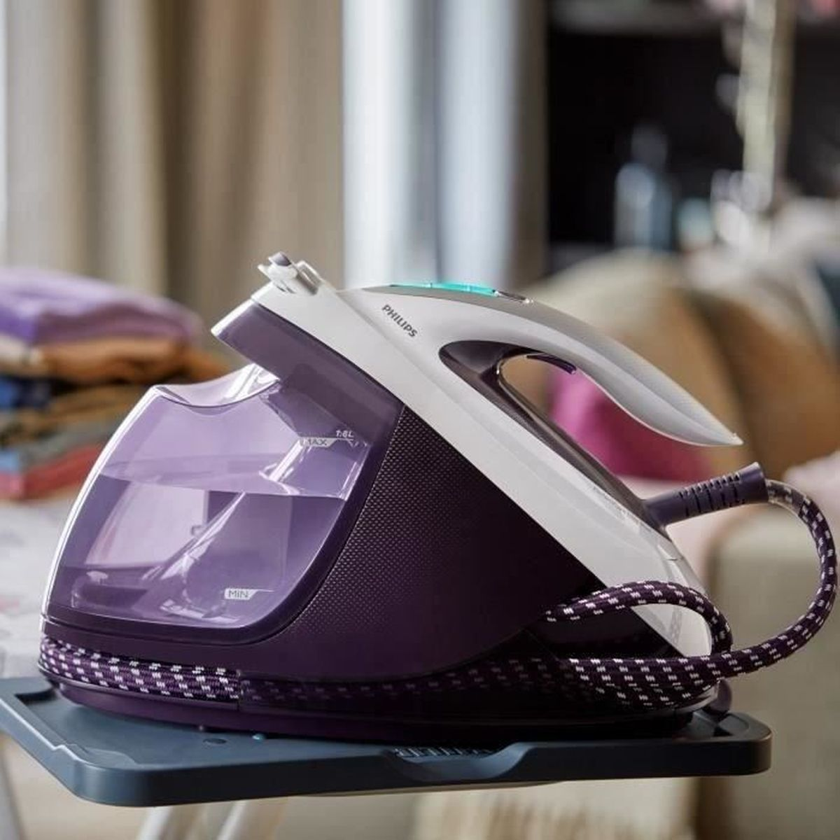 Fioletowo-białe żelazko parowe Philips stoi na tacy. Ma zbiornik na wodę i zwinięty przewód. Tło pokazuje ubrania i zasłonę.