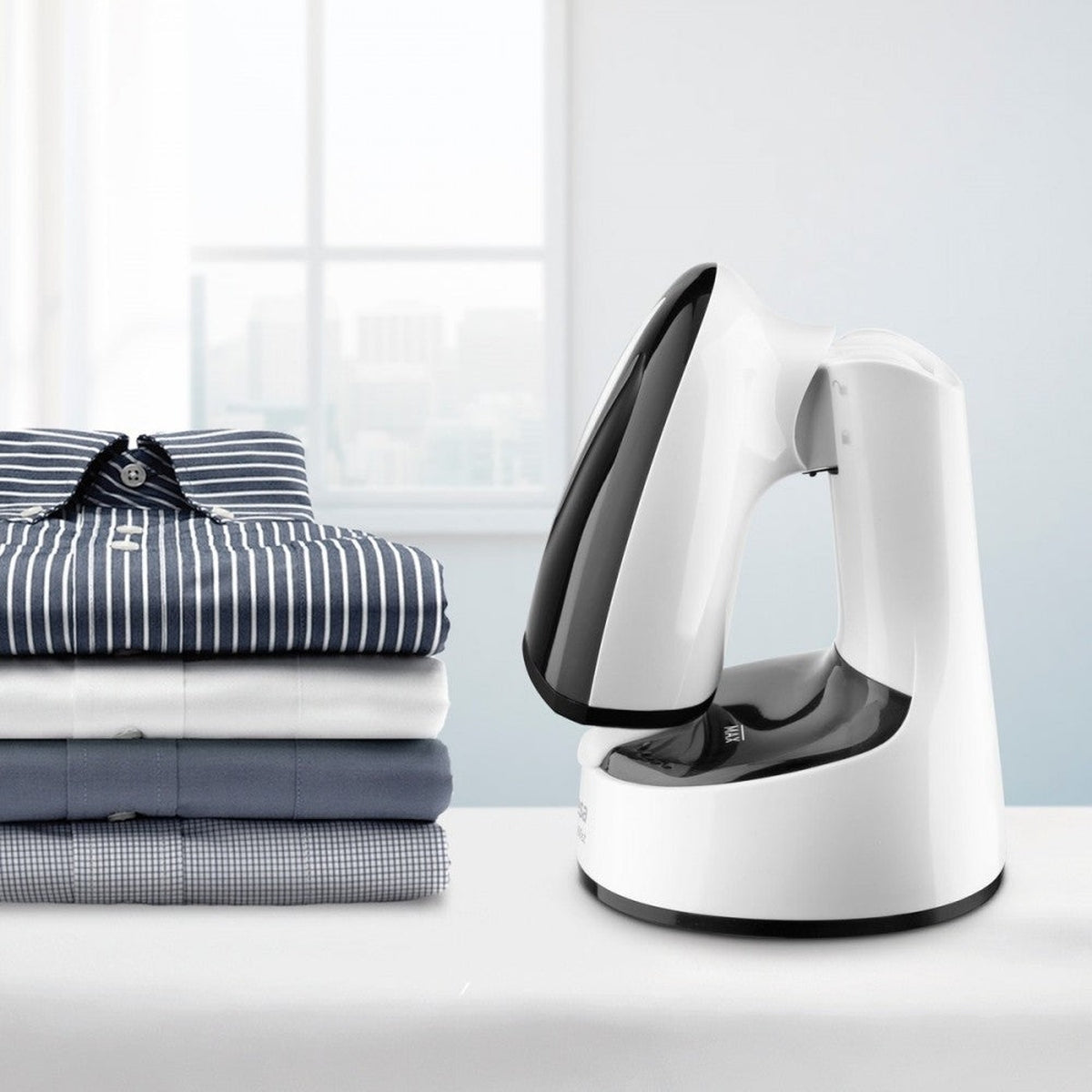 Una pila de camisas dobladas y un vaporizador de ropa blanco y negro sobre una mesa blanca. Hay una ventana en el fondo.