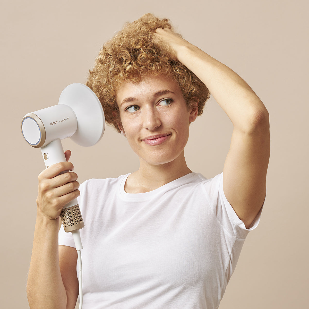 Una mujer con pelo rizado sostiene un secador blanco, tocándose el pelo. Lleva una camiseta blanca, sonriendo. Fondo beige.