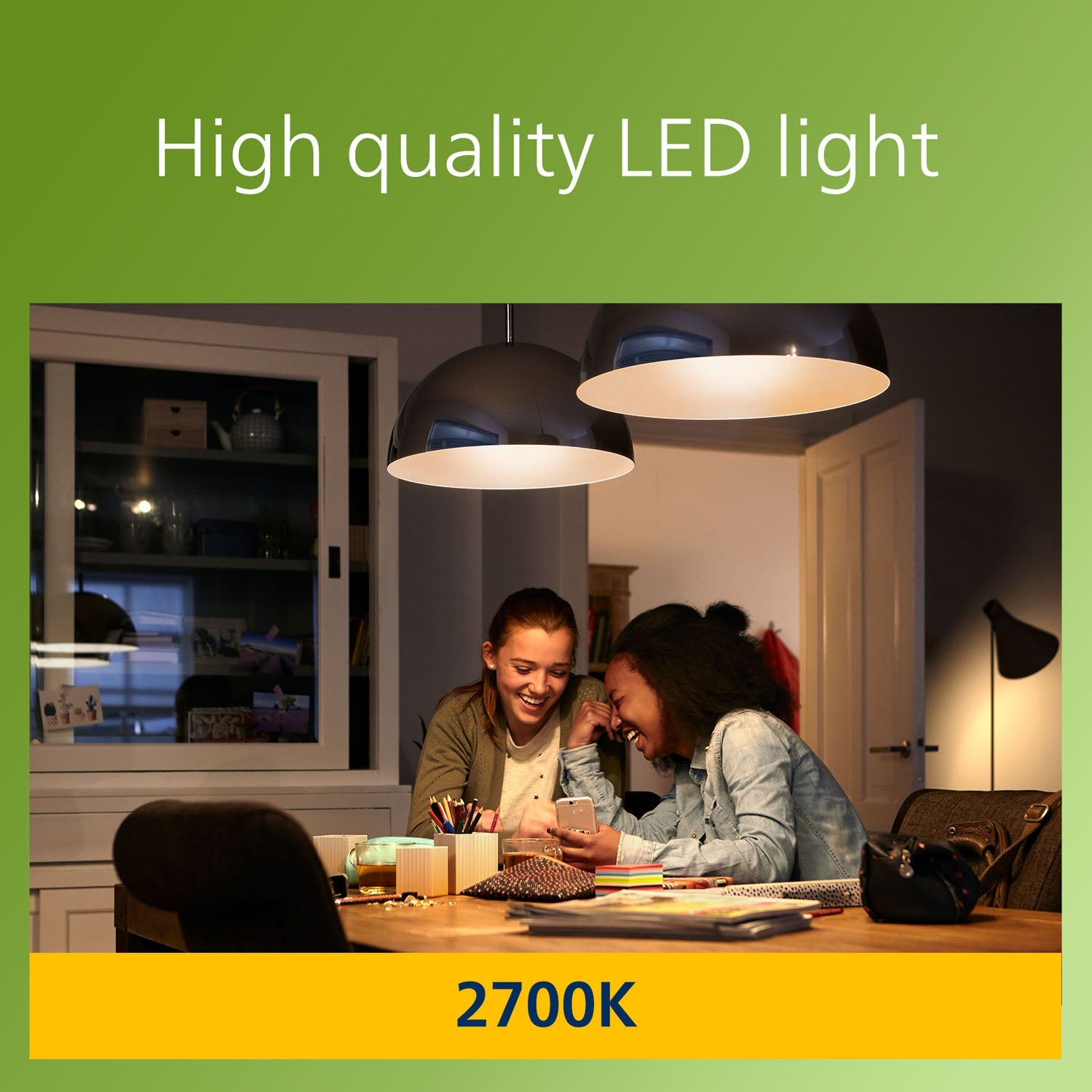 Deux filles souriantes partagent un téléphone, éclairées par des lampes suspendues. Une table en bois est garnie de papeterie. L'arrière-plan est une pièce.