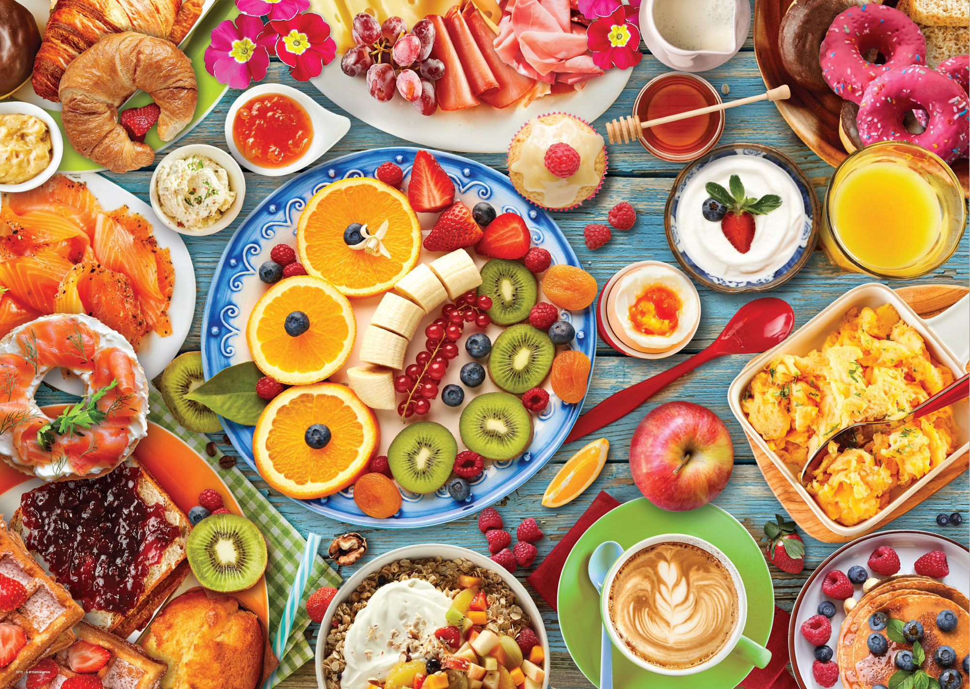 Frühstücksauswahl mit Croissants, Obst, Joghurt, Donuts, Räucherlachs, Eiern, Kaffee und anderen Speisen auf blauem Holztisch.