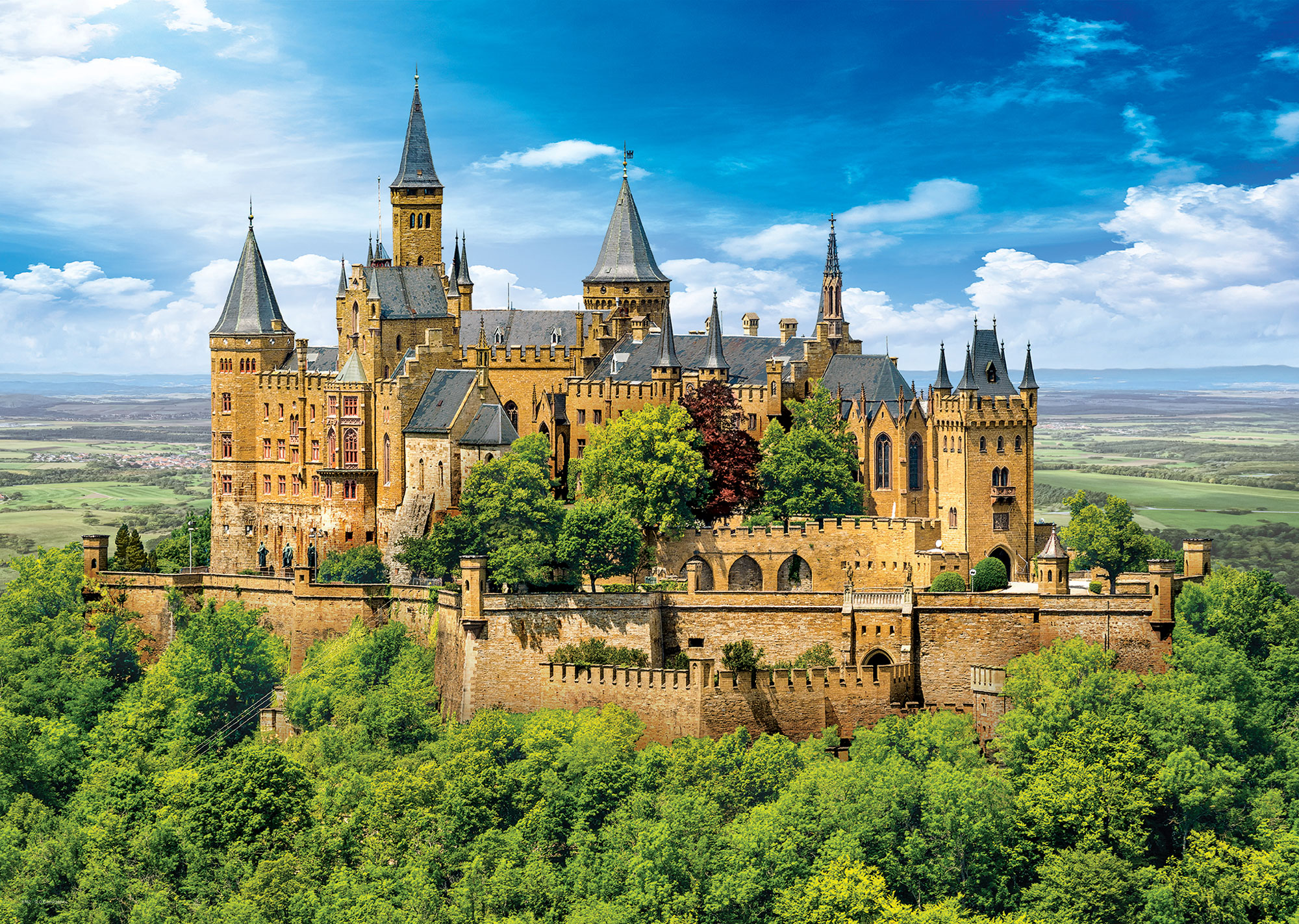 Eine große Burg auf einem Hügel, umgeben von grünen Bäumen, unter blauem Himmel mit Wolken. Die Burg hat mehrere Türme.