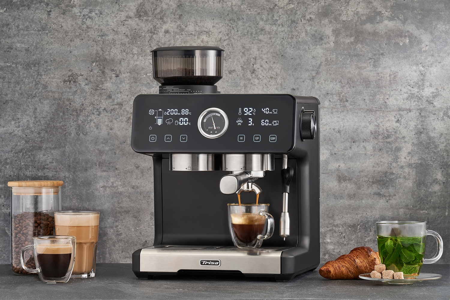 Espressomaschine, die Kaffee in eine Glastasse brüht. Auf dem Tisch befinden sich weitere Getränke, ein Croissant und eine Teetasse.