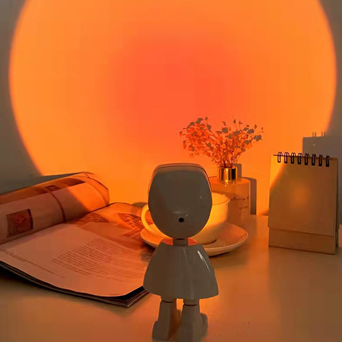 Eine weiße roboterförmige Lampe steht auf einem Tisch. Ein Buch, eine Tasse, Blumen und ein Kalender sind in der Nähe. Orangefarbenes Licht scheint auf die Wand.