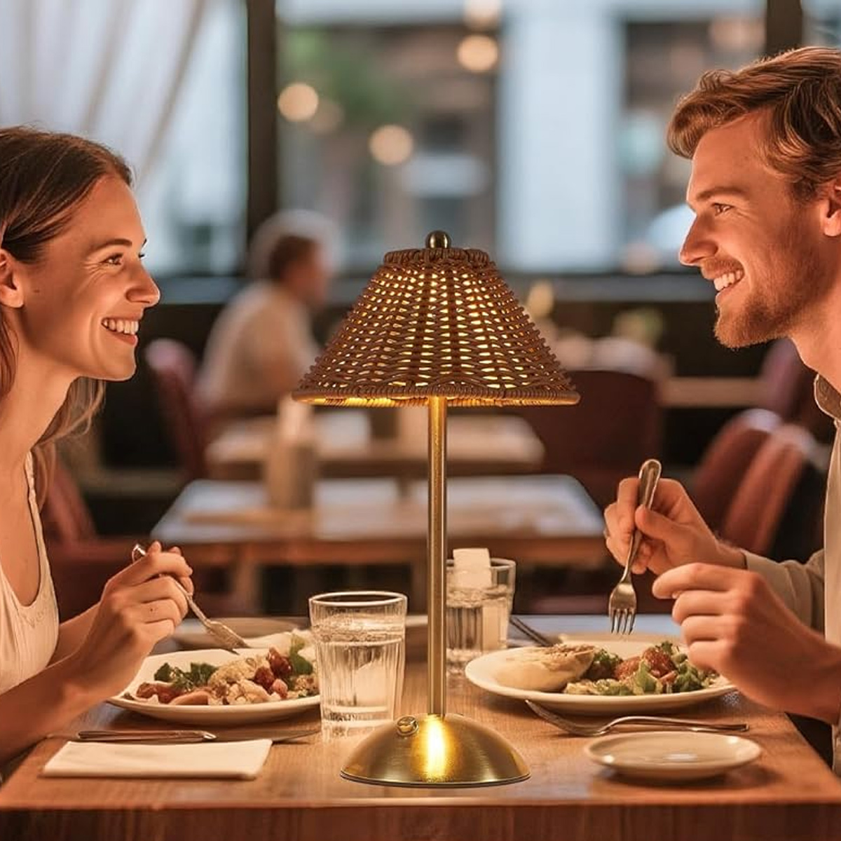 Ein Paar lächelt sich über einen Tisch an. Zwischen ihnen steht eine Lampe. Auf dem Tisch stehen Teller mit Essen, Wassergläser und Besteck.