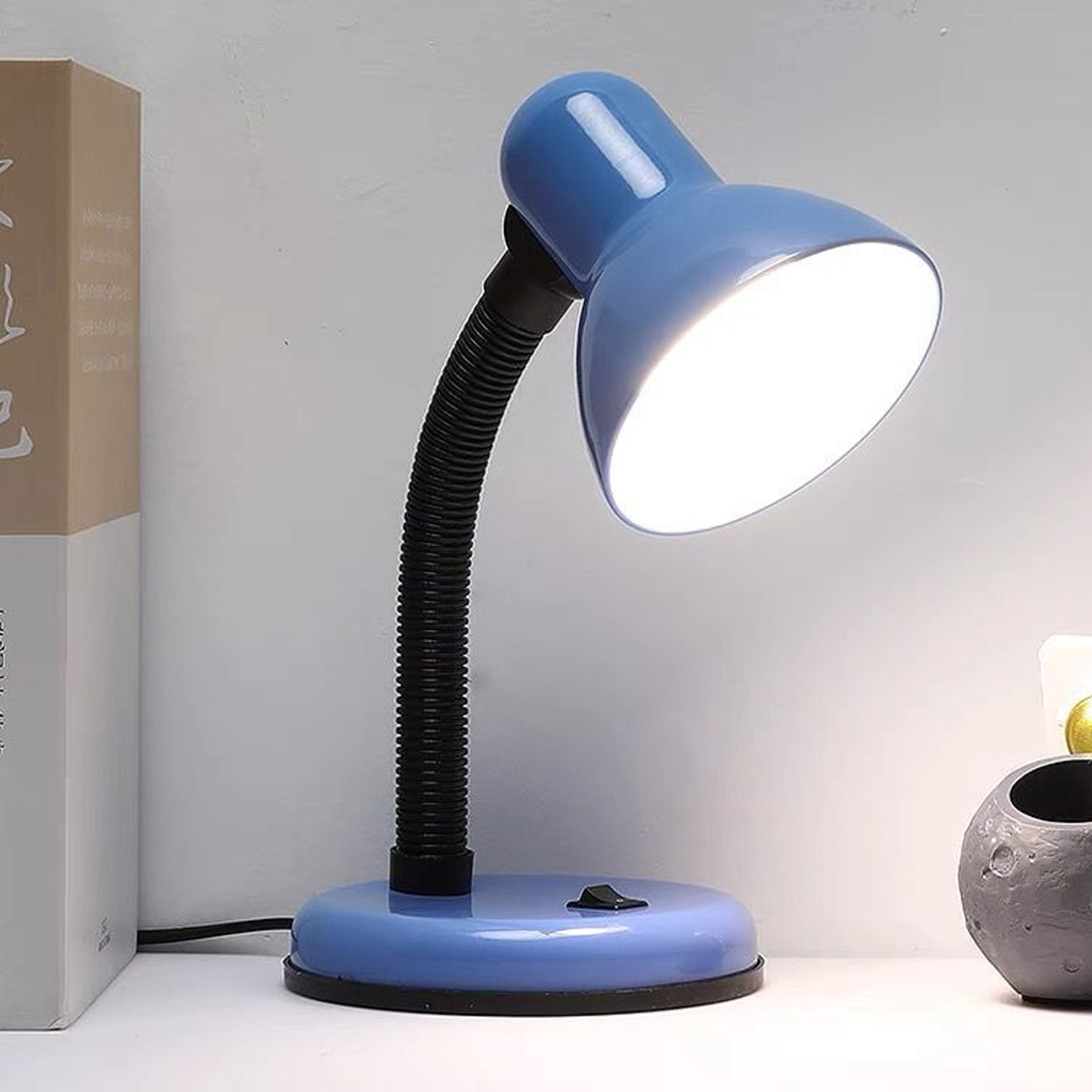 Eine blaue Schreibtischlampe mit schwarzem, flexiblem Hals. Die Lampe ist eingeschaltet und beleuchtet die weiße Oberfläche. Ein Buch und ein mondförmiger Topf sind sichtbar.