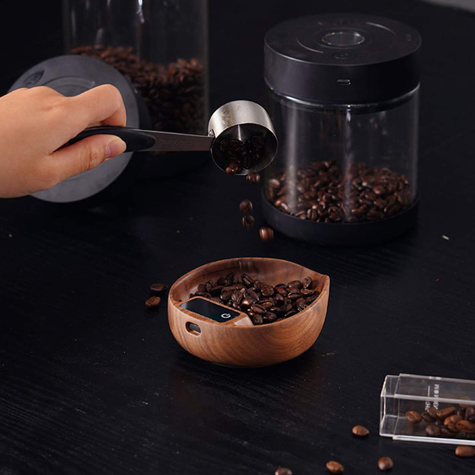 Eine Hand gießt Kaffeebohnen aus einem Löffel in eine Holzschale mit integrierter Waage. Kaffeegläser sind ebenfalls vorhanden.