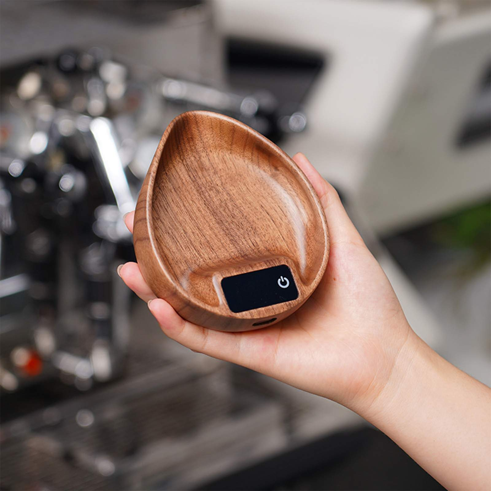 Eine Hand hält eine Holzwaage. Sie hat eine tropfenförmige Form mit einem schwarzen Display und einem Einschalter.