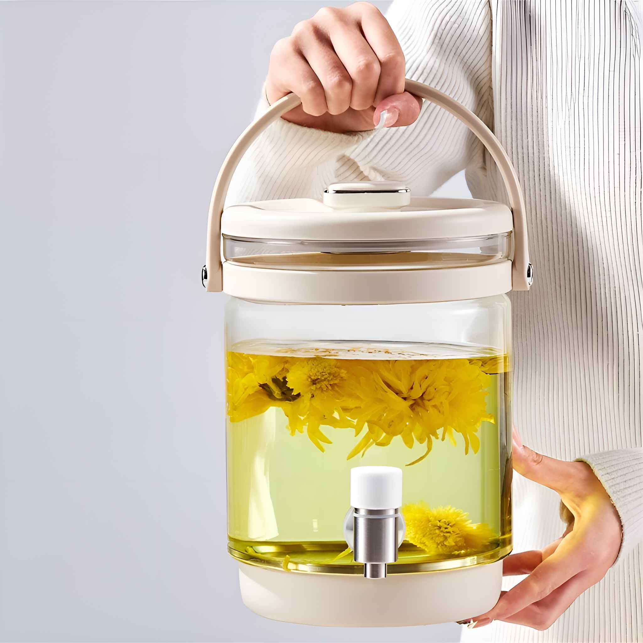Eine Person hält einen Glas-Spender mit gelben Blumen in Wasser. Der Spender hat einen weißen Wasserhahn und Griff.