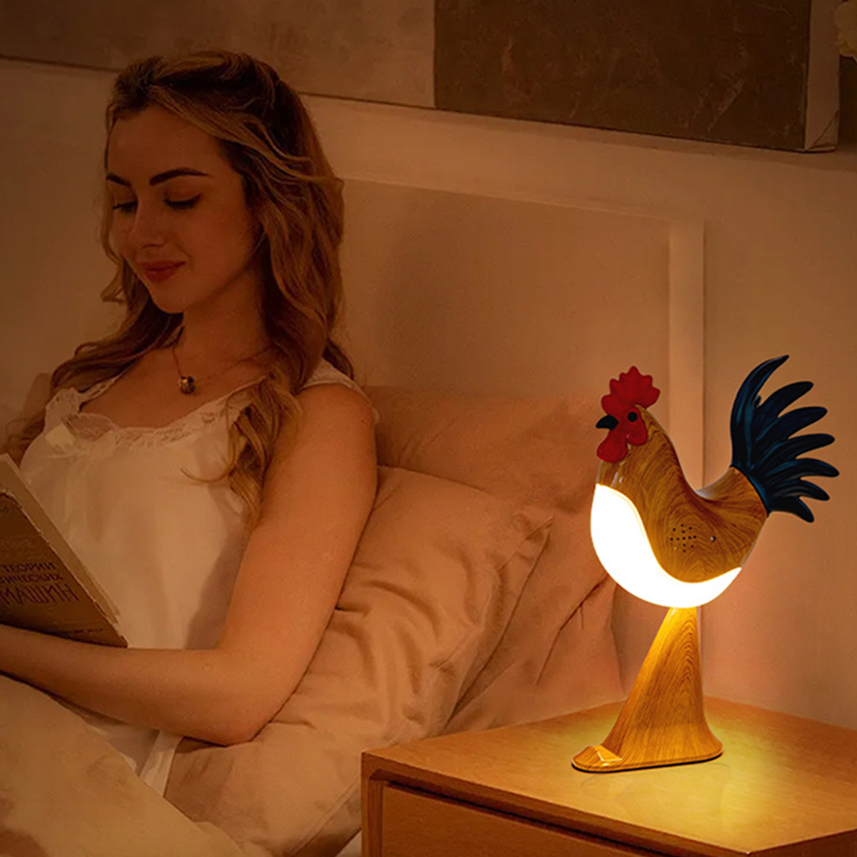Eine Frau liest im Bett, beleuchtet von einer hahnförmigen Lampe auf einem Nachttisch. Die Lampe ist aus Holz mit rotem Kamm und blauem Schwanz.