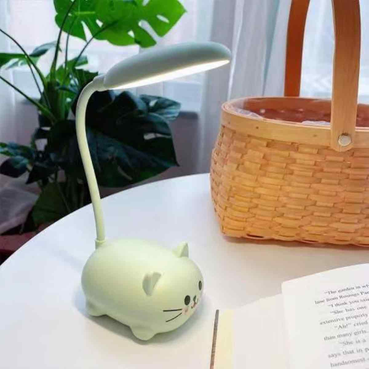 Eine katzenförmige Lampe steht neben einem Korb und einem Buch. Grüne Blätter sind im Hintergrund. Die Lampe ist an.