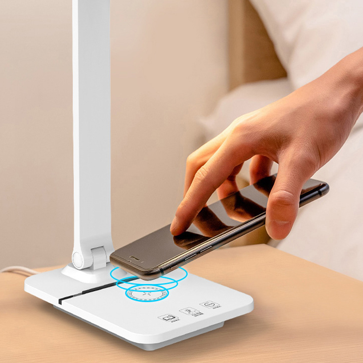 Eine Hand legt ein Smartphone auf ein weißes Ladepad. Daneben steht eine weiße Lampe. Der Hintergrund zeigt ein Bett und einen Nachttisch.
