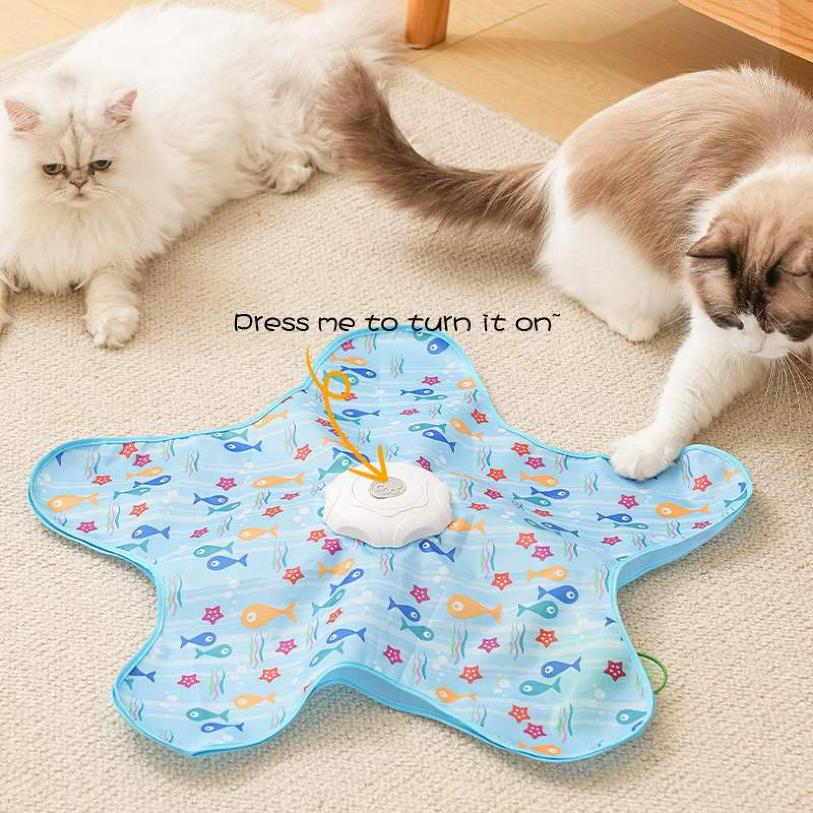 Zwei Katzen in der Nähe einer sternförmigen blauen Matte mit Fischmustern. Die Matte hat einen weißen Knopf. Der Text lautet: 'Dress me to turn it on'.