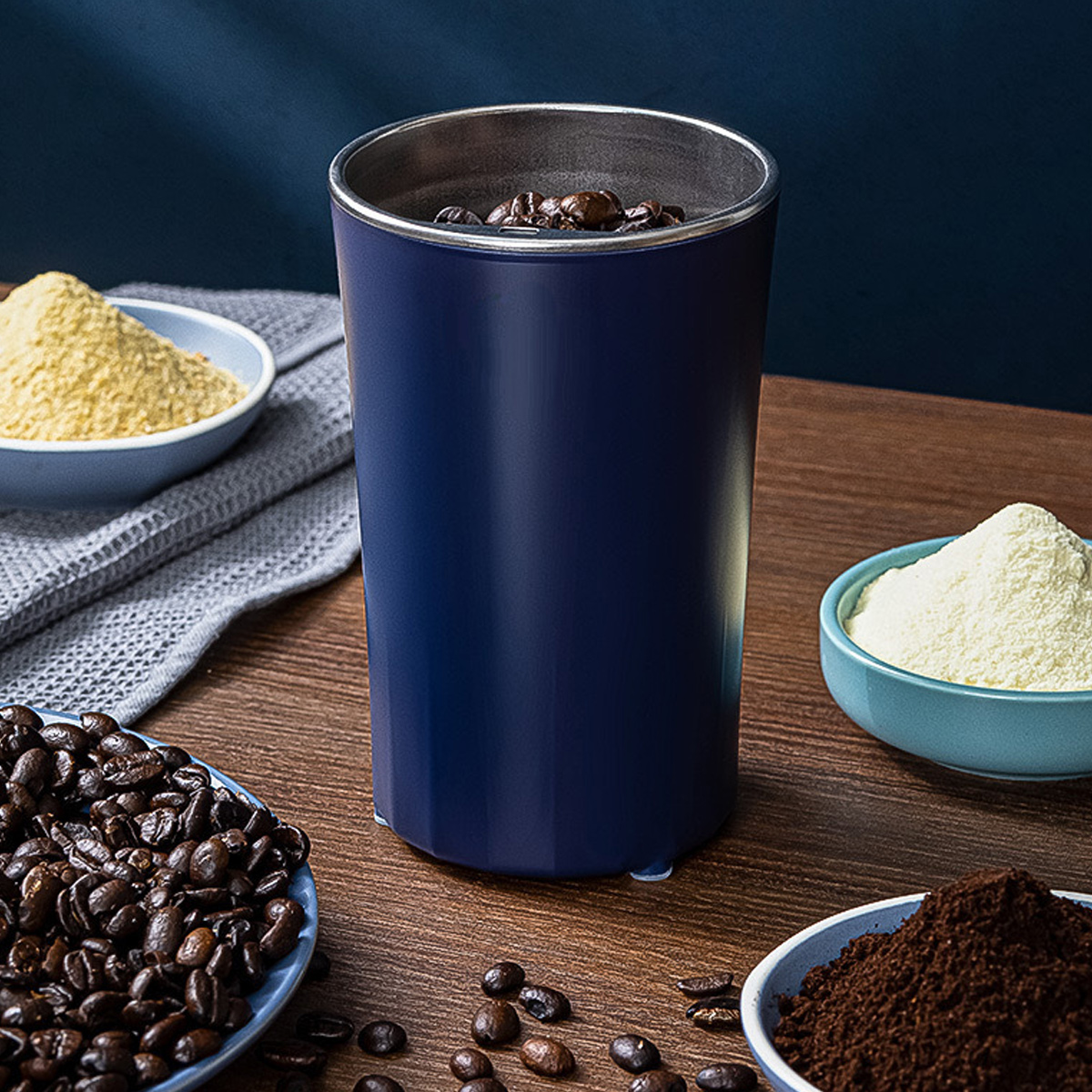 Ein blauer Kaffeemühle mit Bohnen, umgeben von Kaffeebohnen, Pulver und gemahlenem Kaffee in blauen Schalen.